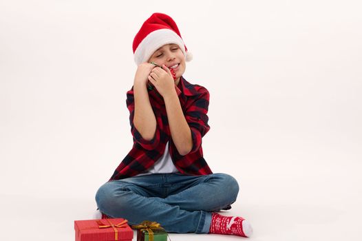 Full length portrait: handsome preteen boy in red plaid shirt and Santa hat, hugging a Christmas gift, sitting on white background with copy space for text. Looking forward to upcoming winter holidays
