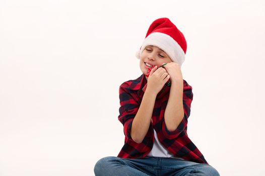 Lovely preteen boy in Santa hat, gently hugging a Christmas gift, isolated over white background, copy advertising space. Boxing Day. Happy festive atmosphere of winter holidays. New Year celebration