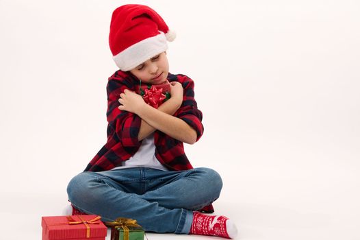 Adorable child boy in Santa hat, gently hugging a Christmas gift, isolated over white background, copy advertising space. Boxing Day. Happy festive atmosphere of winter holidays. New Year celebration