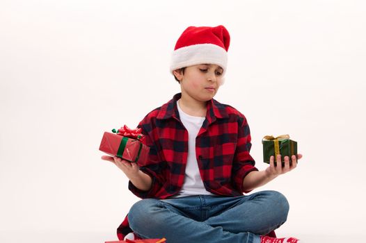 Adorable Caucasian preteen boy wearing Santa hat and red plaid shirt, holding cute Christmas gifts. Isolated portrait on white background. Copy space. Time top open presents. Happy New Year concept