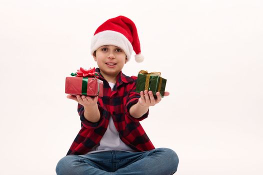 Adorable multi-ethnic preteen boy in Santa hat and red plaid shirt, handing Christmas gifts, looking at camera. Isolated on white background. Copy space. Time top open presents. Happy New Year concept