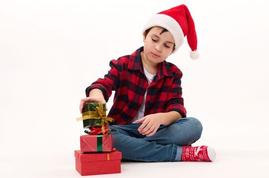 Happy Caucasian preteen boy in Santa hat, red plaid shirt, denim jeans and socks with Christmas pattern, stacks Xmas gifts one on one, isolated on white background. Boxing Day. December 25th. Ad space