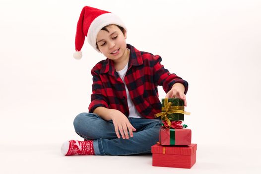 Portrait of a Caucasian lovely preteen boy wearing Santa hat, red plaid shirt, blue denim jeans and socks with Xmas pattern, stacking Christmas gifts, sitting on white background. New Year celebration