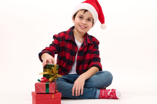 Happy Middle-Eastern preteen boy in Santa hat, red socks with Christmas pattern, smiles looking at camera while folding presents one on one, on a white background. Copy space. Happy Winter holidays