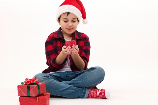 Full length portrait of preteen boy in Santa hat, making wish for Christmas, blowing out red candle. Beautiful gifts lying on white background. Copy space. Looking forward to upcoming winter holidays