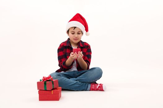 Full length portrait of child boy in Santa hat, making wish for Christmas, blowing out a red candle. Beautiful presents lying on white background. Copy space. Looking forward to upcoming winter events