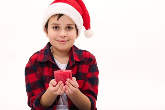 Close-up portrait of an adorable multi-ethnic child boy in Santa hat, cutely smiling looking at camera, holding a lit candle in his hands. Christmas atmosphere. Enjoying the upcoming winter holidays.