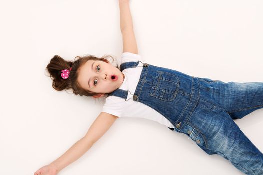 View from above of a happy little child girl, wearing blue denim overalls, experiencing astonishment surprise looking at camera, lying down on a white background with copy advertising space for text