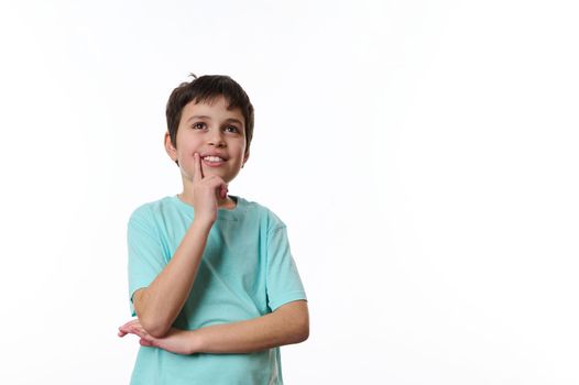 Happy teenage boy wearing turquoise t-shirt, holding his hand at chin and cutely smiling, thoughtfully looking at aside, isolated over white background with copy ad space for promotional text