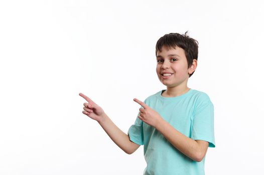 Isolated portrait on white background of a smart school boy, cheerfully smiling teenager wearing blue turquoise t-shirt, looking at you while pointing at advertising space for your promotional text