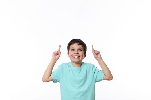 Portrait of Caucasian handsome pleasant pre-adolescent boy in blue casual t-shirt, smiling pleasantly, looking up, pointing finger at copy space for your advertising text, isolated on white background