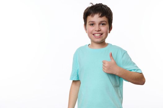 Happy Caucasian preteen child boy in blue t-shirt, a smart schoolboy shows a thumb up, cutely smiles looking at camera, isolated over white background. Free advertising space for your promotional text