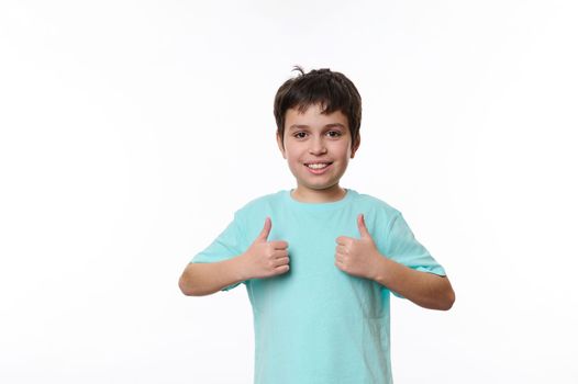 Portrait of Caucasian handsome pleasant teenage child boy in blue casual t-shirt, smiling pleasantly, looking at camera, showing thumbs up, isolated on white background with space for promotional text