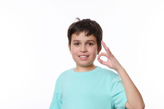 Dark-haired Caucasian boy in blue turquoise casual t-shirt, smiling a cheerful beautiful toothy smile, showing ok sign looking at camera, isolated over white background with space for promotional text