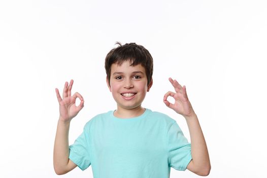 Happy Caucasian preteen child boy in blue t-shirt, a smart schoolboy shows OK sign, cutely smiles looking at camera, isolated over white background. Free advertising space for your promotional text