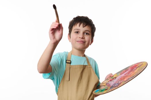 Portrait on white background of talented handsome teenage boy, artist painter holding a palette of paints, painting with paintbrush in front of camera. Creative art development and kids entertainment