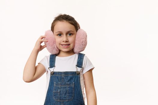 Happy child, lovely baby girl wearing cute pink winter earmuffs - plush headphones, cutely smiling looking at camera, isolated over white background. Protect your ears from cold. Copy space for text