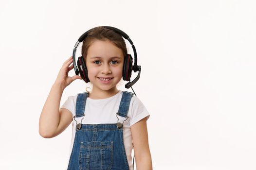 Beautiful smart little school girl, first grader wearing audio headset, ready for online lesson, smiling cutely looking at camera, isolated over white background with copy advertising space for text