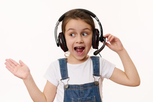 Caucasian smart little child, a school girl wearing audio headset, looking aside, holding imaginary copy space on her hand palm up, on white background. Online education, communication via video link