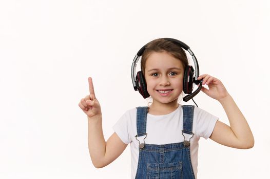 Adorable little girl, smart child first grader wearing headphones, cutely smiles looking at camera, pointing at a copy space on white background. Online education. Distance learning. Video call