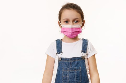 Beautiful European little girl in blue denim overalls, wearing a pink protective mask, isolated over white background. Free space for your advertising text. Pediatrics. Health care and medical content