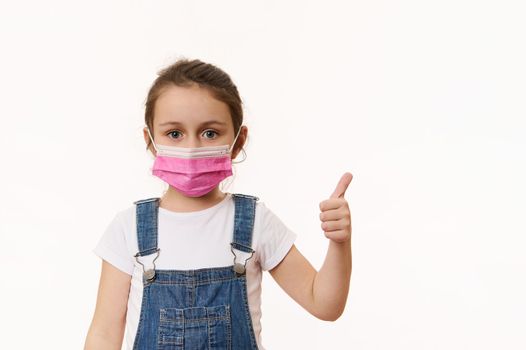 Cute child girl in blue denim overalls, wearing a pink safety mask, shows thumb up, looking at camera, isolated over white background. Space for promo text. Pediatrics. Health care and medical content