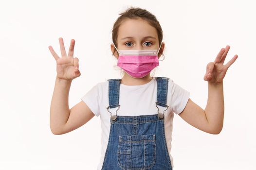 Adorable Caucasian little girl wearing pink protective medical mask, gestures with hands, shows OK signs, looks at camera, isolated over white background. Free space. Pediatrics. Health care. Medicine