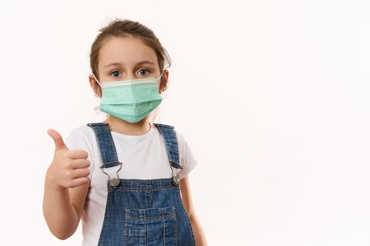 Lovely little child girl in blue denim overalls, wearing green medical protectcive mask, shows thumb up, looking at camera. White background copy space. Stay at home. Pediatrics. Healthcare. Medicine