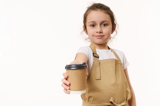 Caucasian lovely little girl wearing a beige chef's apron, handing out on camera a takeaway hot tea coffee drink in cardboard mug, isolated on white, copy space for text. Beverage in paper cup to go
