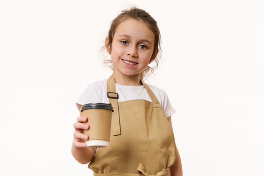 Beautiful blue-eyed Caucasian little girl wearing a beige chef's apron, handing out a takeaway hot beverage, tea or coffee drink in an ecologically-friendly paper cup, cutely smiles looking at camera