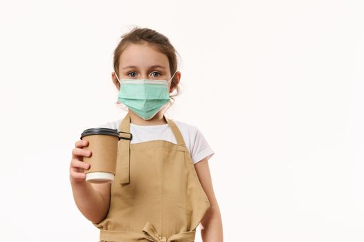 Beautiful blue-eyed European little child girl wearing a beige chef's apron, handing out a takeaway hot beverage, tea coffee drink in ecologically-friendly paper cup, cutely smiles looking at camera