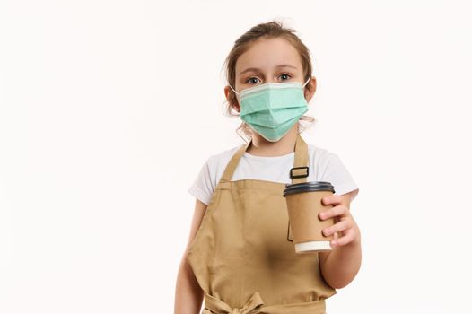 Caucasian little child girl wearing safety medical mask, dressed as barista, bartender with a hot drink in paper cup, isolated on white background. Cafe restaurant on pandemic lockdown. Takeaway food.