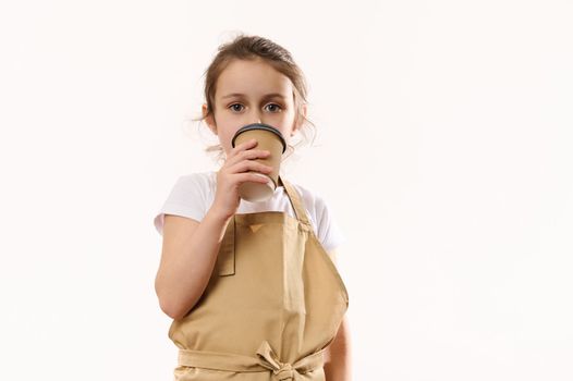 Caucasian beautiful little child girl dressed as barista bartender in beige chef's apron, drinks hot drink from an ecologically-friendly paper cup to go, smiling looking at camera, isolated on white