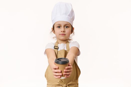 Isolated portrait on white background of cute baby girl wearing a white chef's hat and beige apron, holding, selling a hot drink in a paper cup to go, looking at camera, isolated over white background
