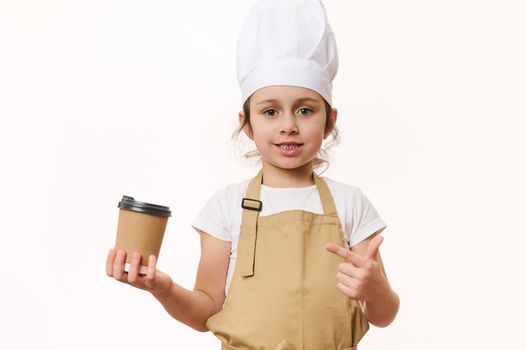 Cute baby girl - barista bartender in white chef hat and beige apron, smiling a beautiful toothy smile looking at camera, pointing at a hot drink in eco paper cup to go, isolated on white background
