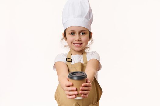 Caucasian little girl dressed as a barista, bartender in white chef hat and apron, smiling a beautiful toothy smile looking and handing on camera a hot drink in eco paper cup to go, isolated on white