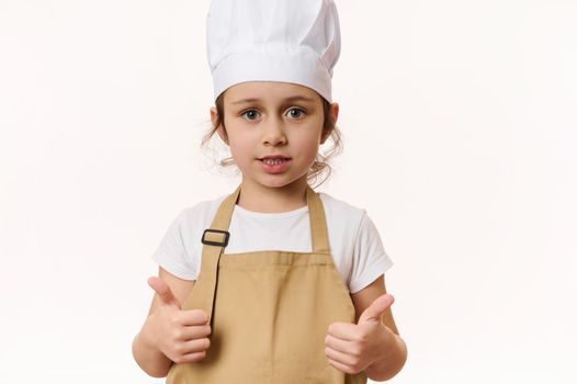 Isolated portrait on white background of adorable Caucasian child, lovely little girl - baker confectioner, cook wearing chef's hat and beige apron, shows thumbs up, looking at camera. Free ad space