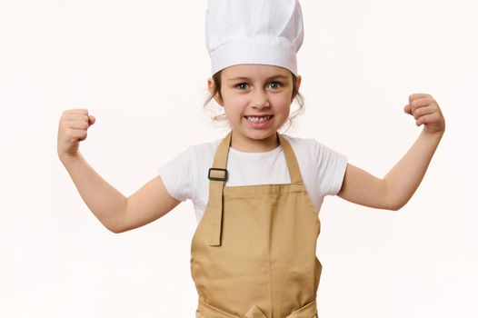 Delighted child, lovely baby girl dressed as pastry chef or baker confectioner, in hat and apron, clenching fists, looking at camera, isolated on white background with free space for advertising text