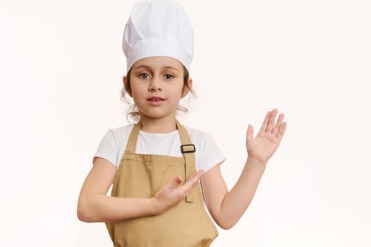 Delighted baby girl dressed as pastry chef or baker confectioner, in chef's hat and apron, pointing at copy space for your promotional text on white background. Concept of bakery, culinary, cookery