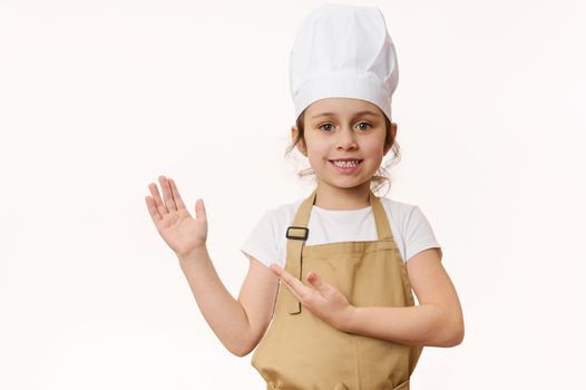 Charming little girl in chef's uniform, smiling sweetly at camera, pointing her hand at a blank place for advertising text, isolated on a white background. Concept for cooking class. Food consumerism