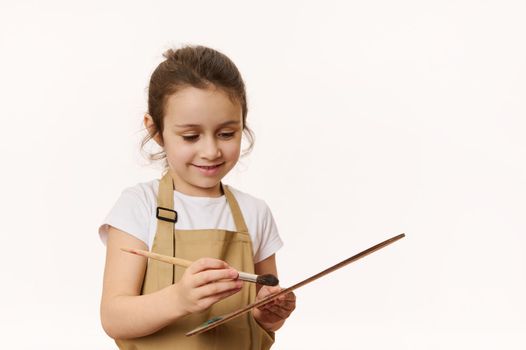 Inspired talented Caucasian little girl painter, artist wearing a beige apron, dipping a paintbrush on wooden palette with colorful oil paints, isolated on white background. Copy advertising space.