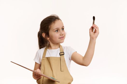 Inspired little child artist, talented baby girl wearing beige apron, with a wooden palette of colorful oil paints and paintbrush, isolated over white background. Art. Creativity. Kids development