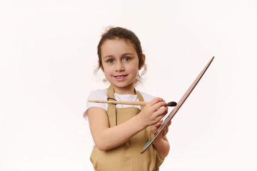 Caucasian lovely baby girl in beige apron, a talented little artist with a paintbrush and palette of colorful paints, smiling a beautiful toothy smile looking at camera, isolated on white background