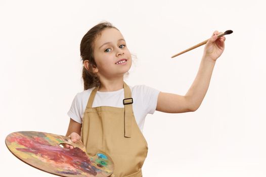Lovely baby girl in beige apron, talented little painter with a paintbrush and palette of colorful paints, cutely smiling a toothy smile looking at camera, white background. Kids art development