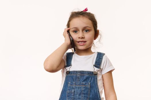 Adorable Caucasian little girl, wearing blue denim overalls and white t-shirt, talking on mobile phone, isolated over white background. Copy advertising space. Kids and electronic digital technology