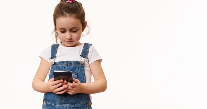 Isolated portrait on white background of a lovely little preschool girl, first grader with a smartphone. Adorable child using mobile phone, checks contents, makes call. Kids and digital technologies
