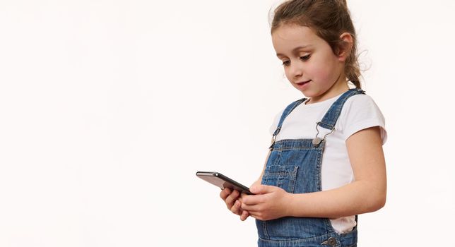 Studio shot on white background: Lovely child little girl using smartphone, plays game on mobile device, pleasantly surprised by easy application. Wow, great app for kids. Internet and digital gadgets