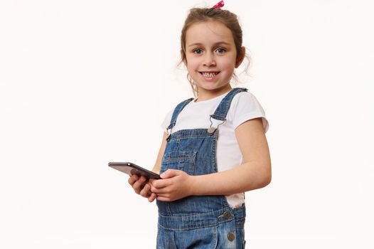 Adorable overjoyed little girl wearing blue denim overalls, holds a modern smartphone in her hands, cutely smiles looking at camera, posing isolated over white background with copy advertising space