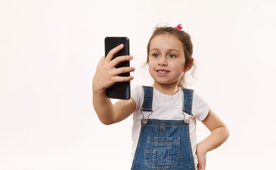 Happy adorable Caucasian little child girl using mobile phone, talks with her parent via video link, isolated over white background with copy space for advertising text. Online chat on smartphone