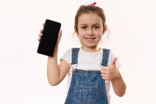 Lovely little kid, cute baby girl holding smartphone with blank screen with advertising free space for promo text, smiling with toothy smile, showing thumb at camera. Children and digital gadgets